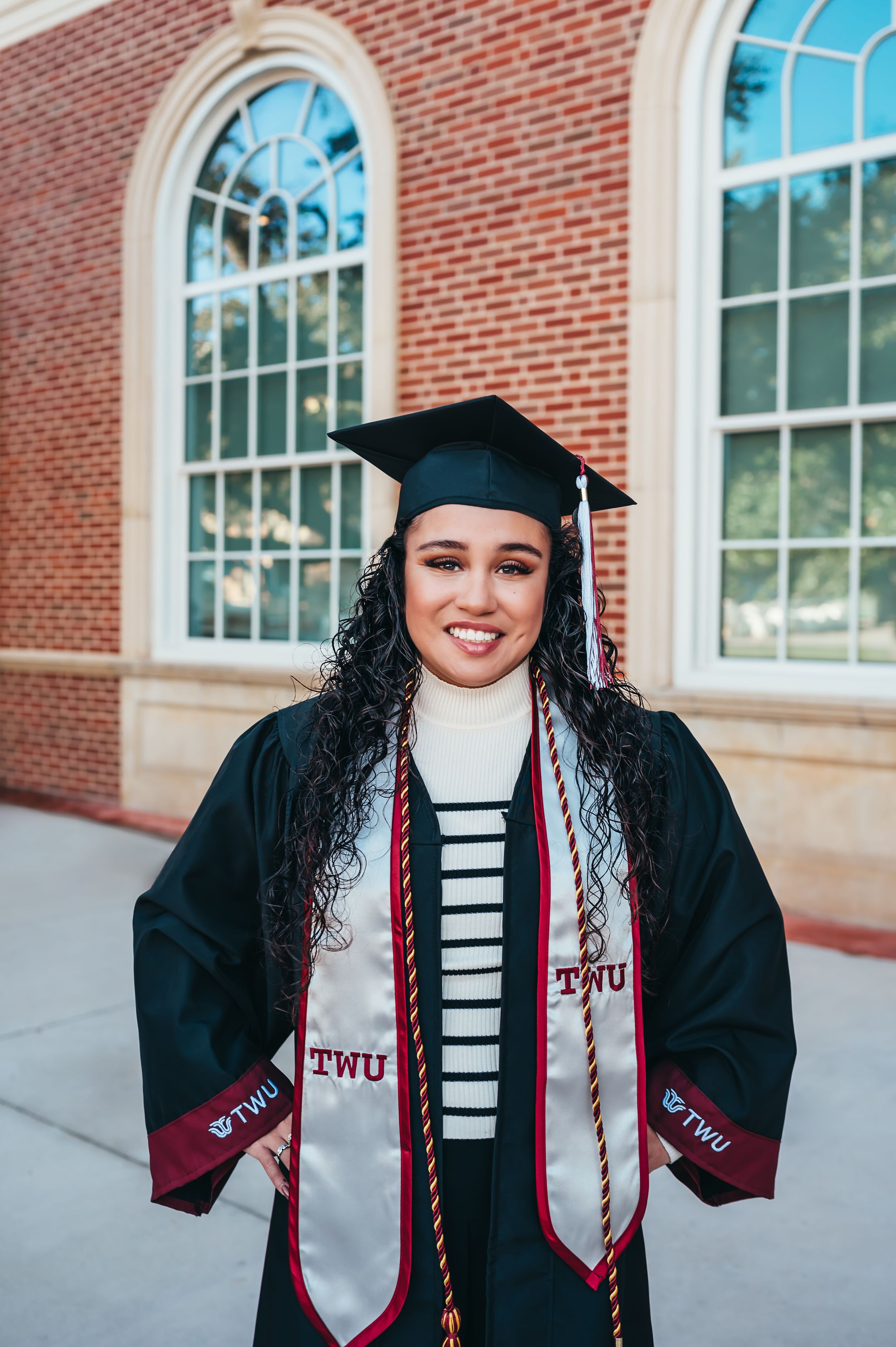 Smiling graduate in cap and gown with TWU stole standing in front of a brick building.