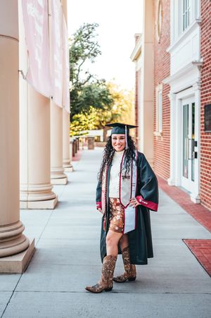 Graduate in cap and gown with cowboy boots posing between columns on a college campus walkway.