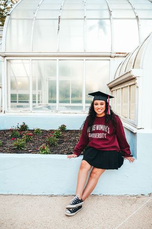 Smiling graduate in Texas Womans University sweatshirt and cap sitting outside greenhouse.