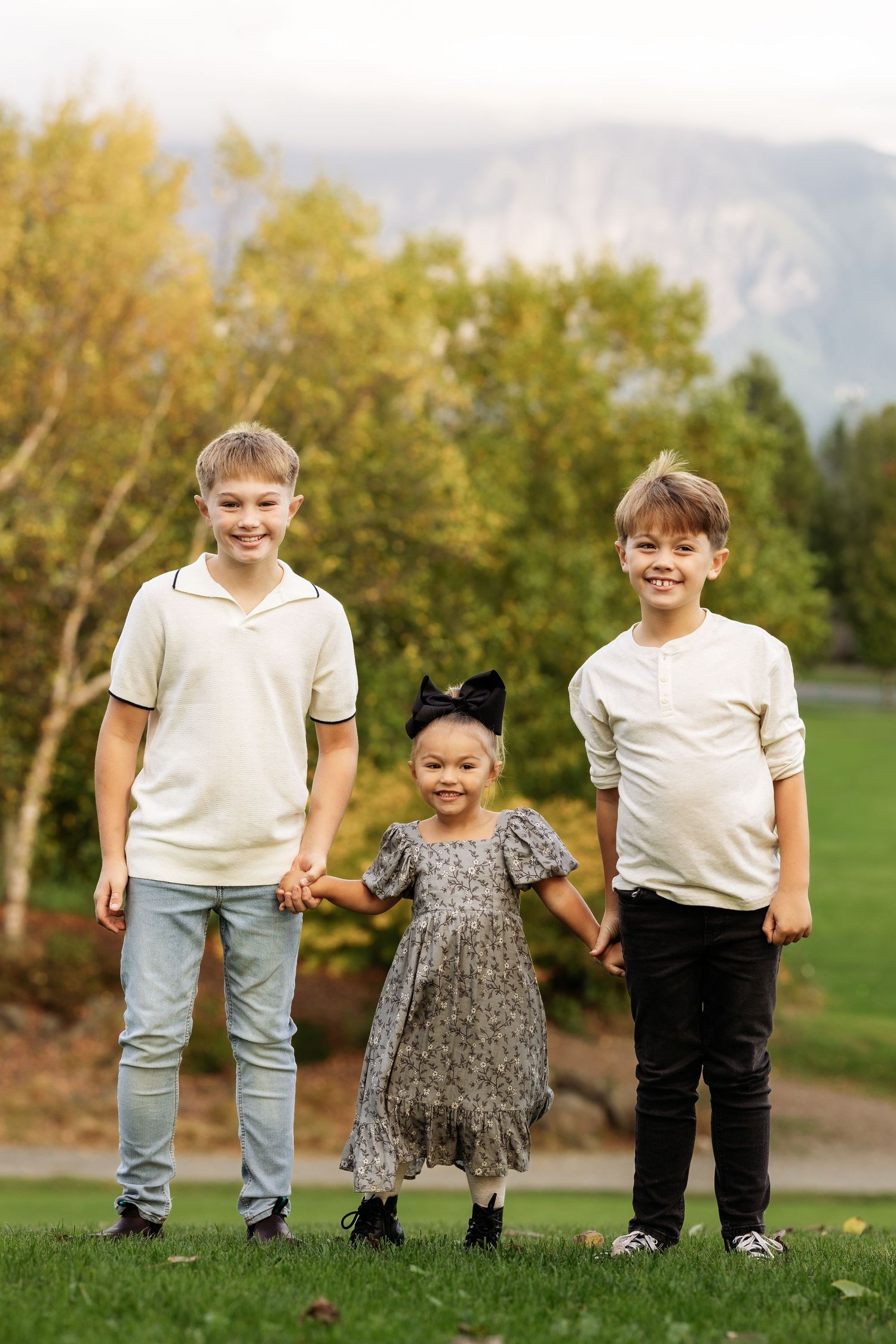 Two smiling boys in cream tops hold hands with a little girl in a floral dress outdoors with mountains behind.