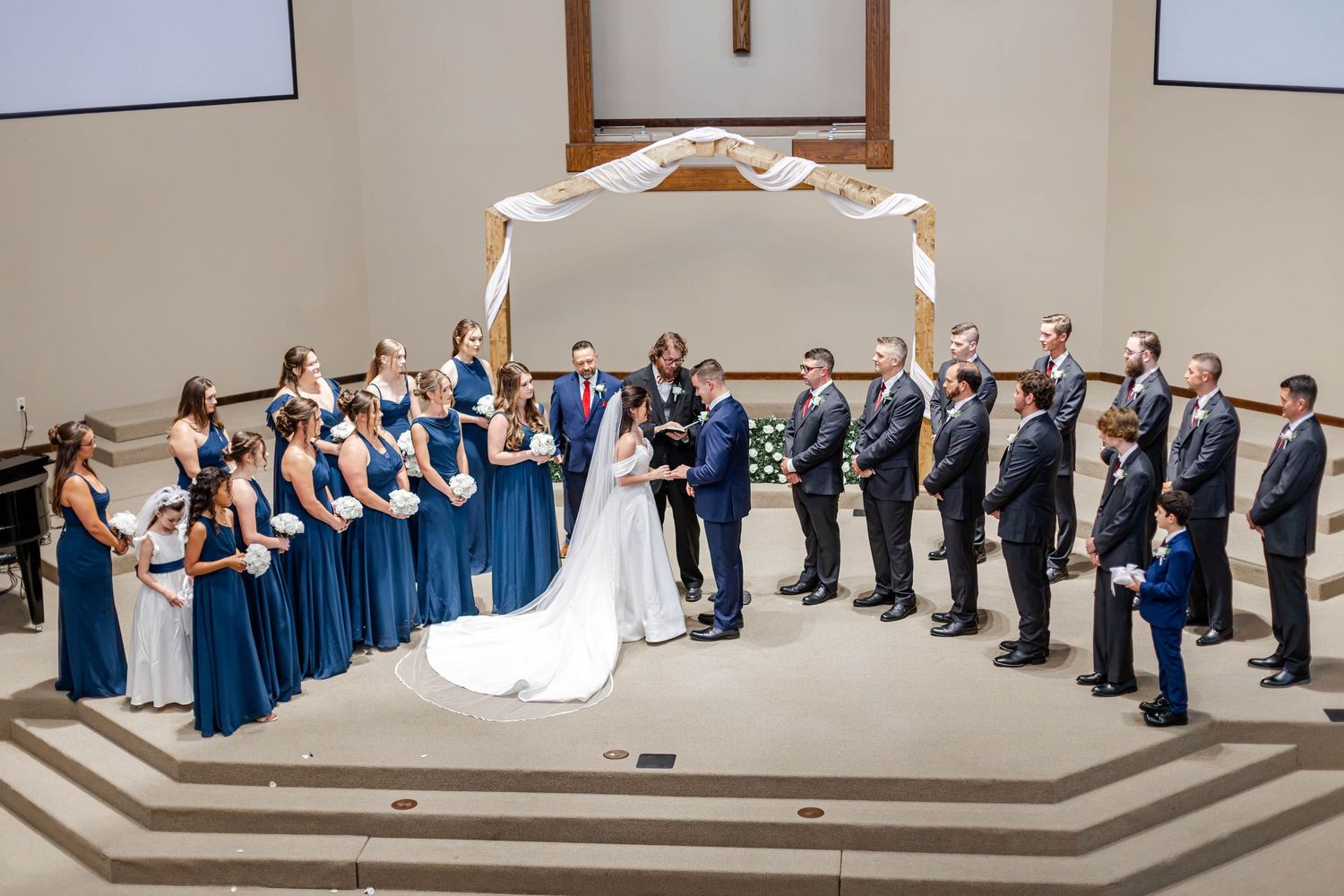 Wedding ceremony in a church with bridesmaids in blue dresses and groomsmen in suits standing beneath a floral arch.