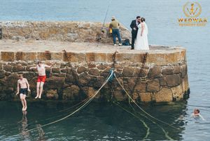 Wedding photo with people swimming and jumping off stone pier by the ocean.