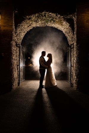 Romantic silhouette of couple under floral arch with dramatic backlighting and fog effects.