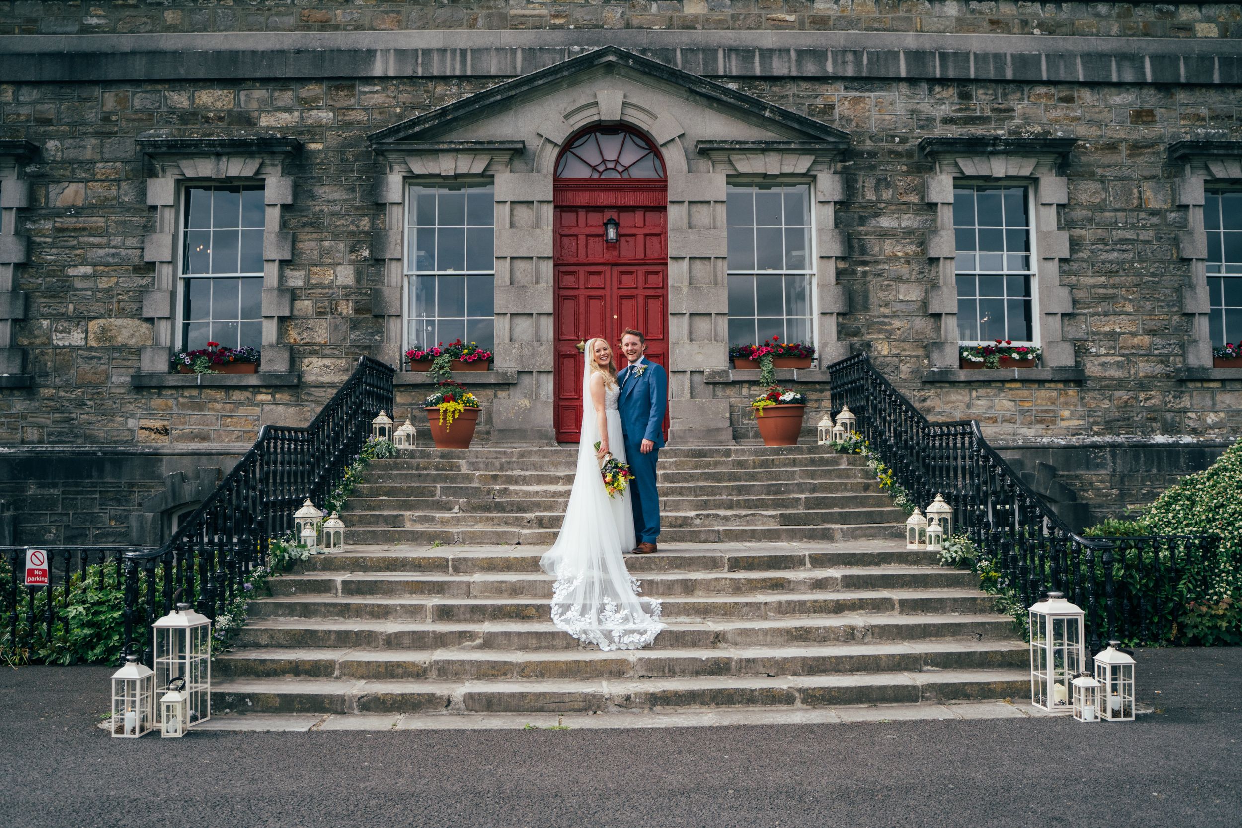 Elegant wedding couple stands on stone steps of historic building with red door and lantern decorations Bellinter House.