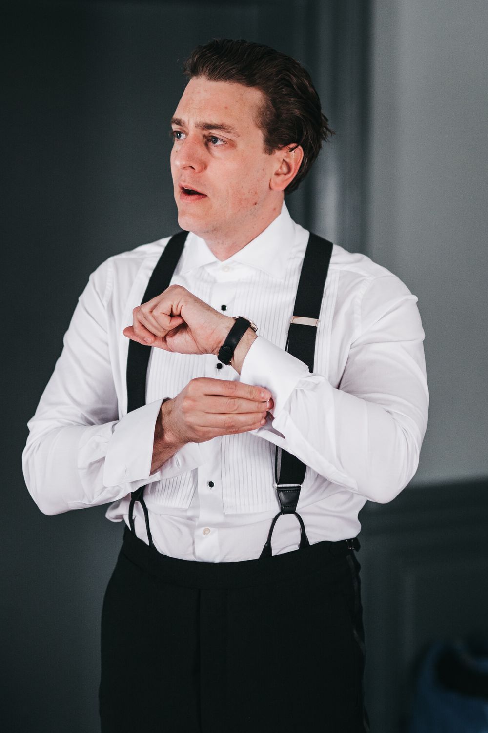 Man in white dress shirt and black suspenders adjusting his cufflinks, getting ready for a formal event.