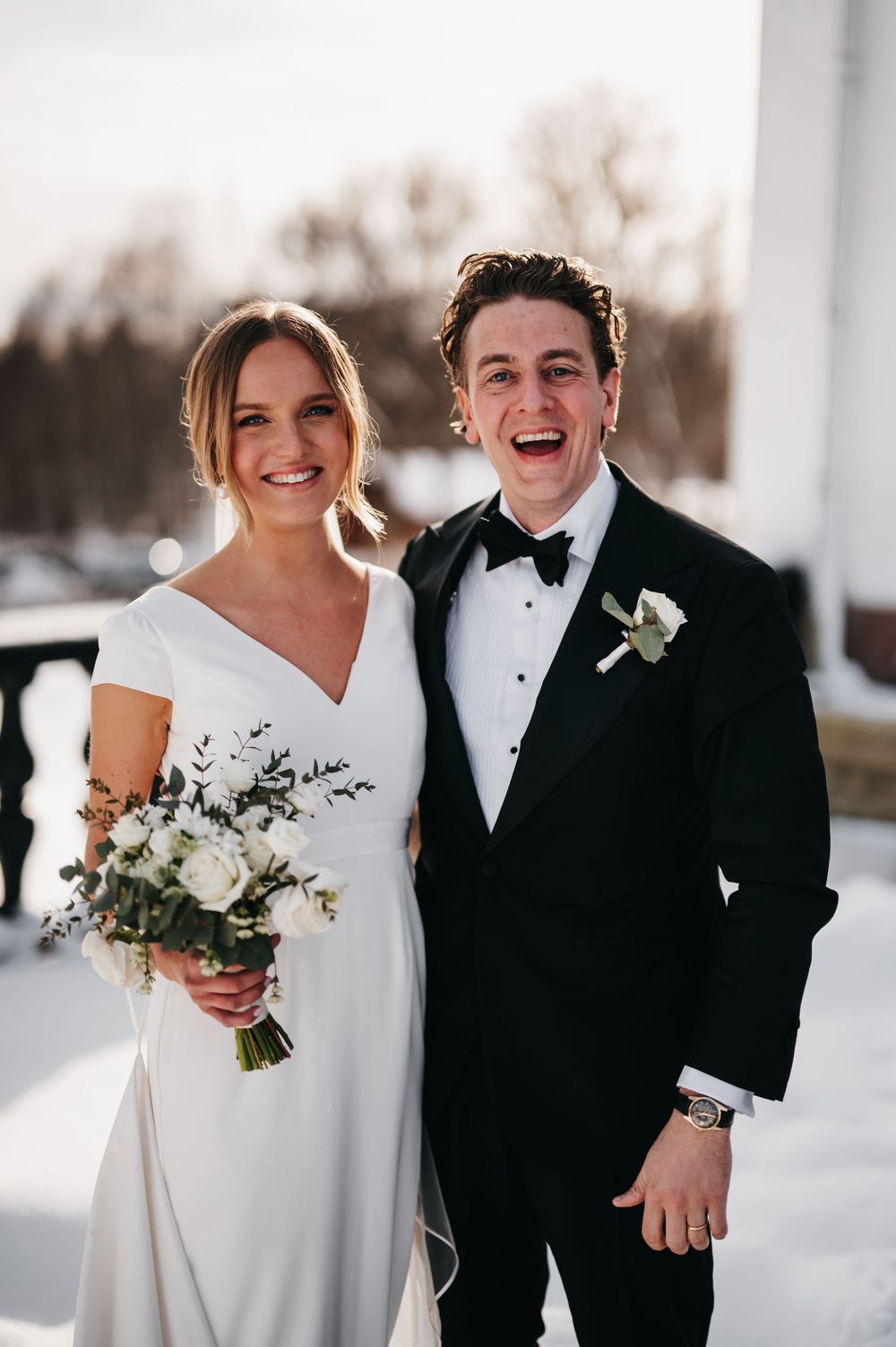 A smiling bride in a white gown holding a bouquet stands beside a laughing groom in a black tuxedo outdoors in winter.