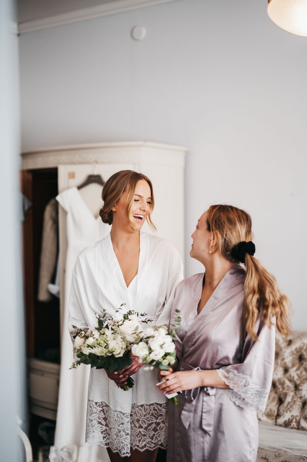 Bride in white lace robe laughing with bridesmaid in lavender robe holding white floral bouquet during wedding preparations.