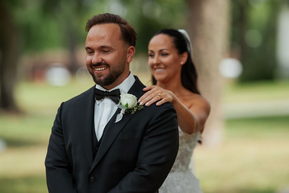 Bride in white gown places hand on groom's shoulder in black tuxedo during outdoor wedding first look.