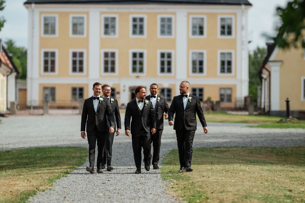 Five groomsmen in black tuxedos walking confidently on a gravel path in front of a elegant yellow manor house.