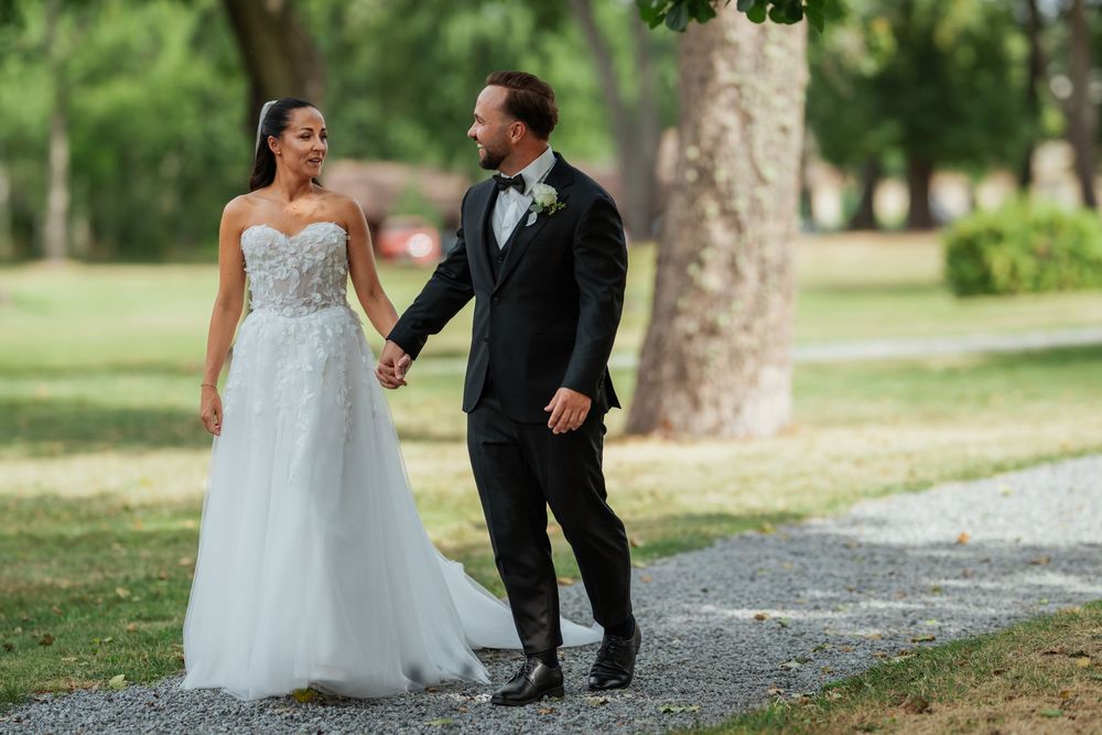 A bride and groom walk hand in hand along a tree-lined path, smiling at each other on their wedding day.