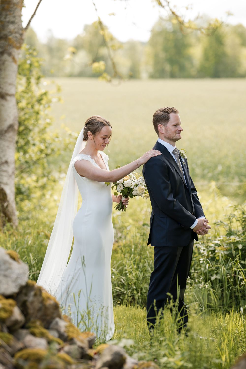 Bride in white gown touches groom's shoulder during outdoor first look at scenic green field wedding.