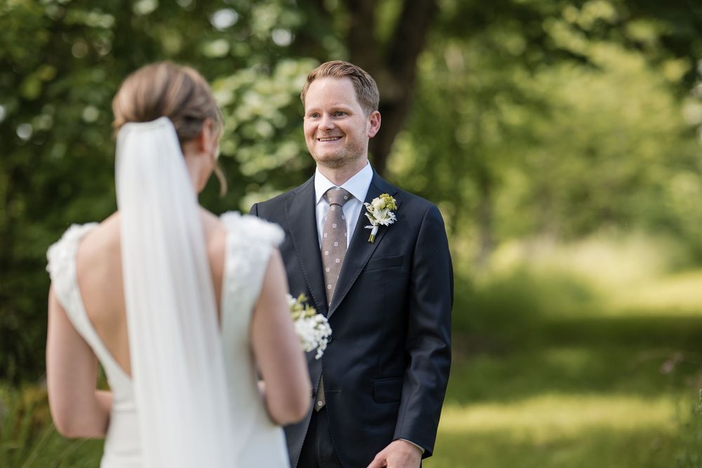 Groom in navy suit smiling at bride in white wedding dress and veil during outdoor first look ceremony.