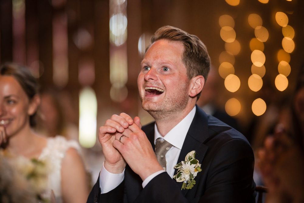 Smiling groom in dark suit with boutonniere clapping joyfully at wedding reception with bokeh lights.