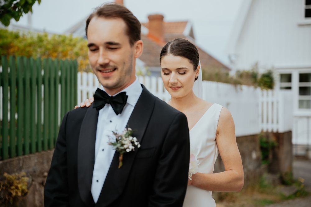 Bride and groom in elegant wedding attire pose outdoors, groom in black tuxedo with boutonniere, bride in white gown.