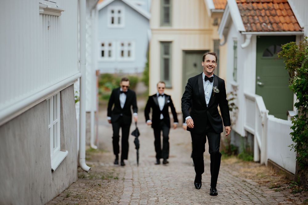 Groom and groomsmen in black tuxedos walking down a cobblestone alley between white Scandinavian houses.