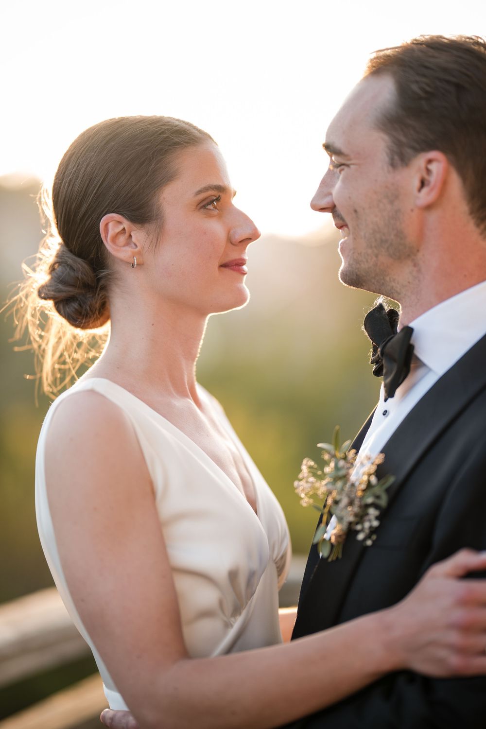 Bride and groom gazing at each other during golden hour, bride in white gown, groom in tuxedo with boutonniere.