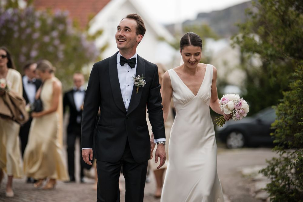 A smiling bride in a white satin gown holds a bouquet beside a groom in a black tuxedo at an outdoor wedding.
