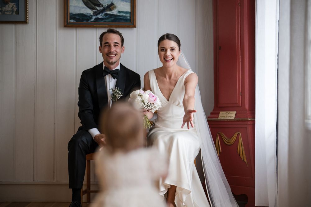 Bride and groom laughing joyfully while seated, with a flower girl in foreground, during elegant wedding ceremony.