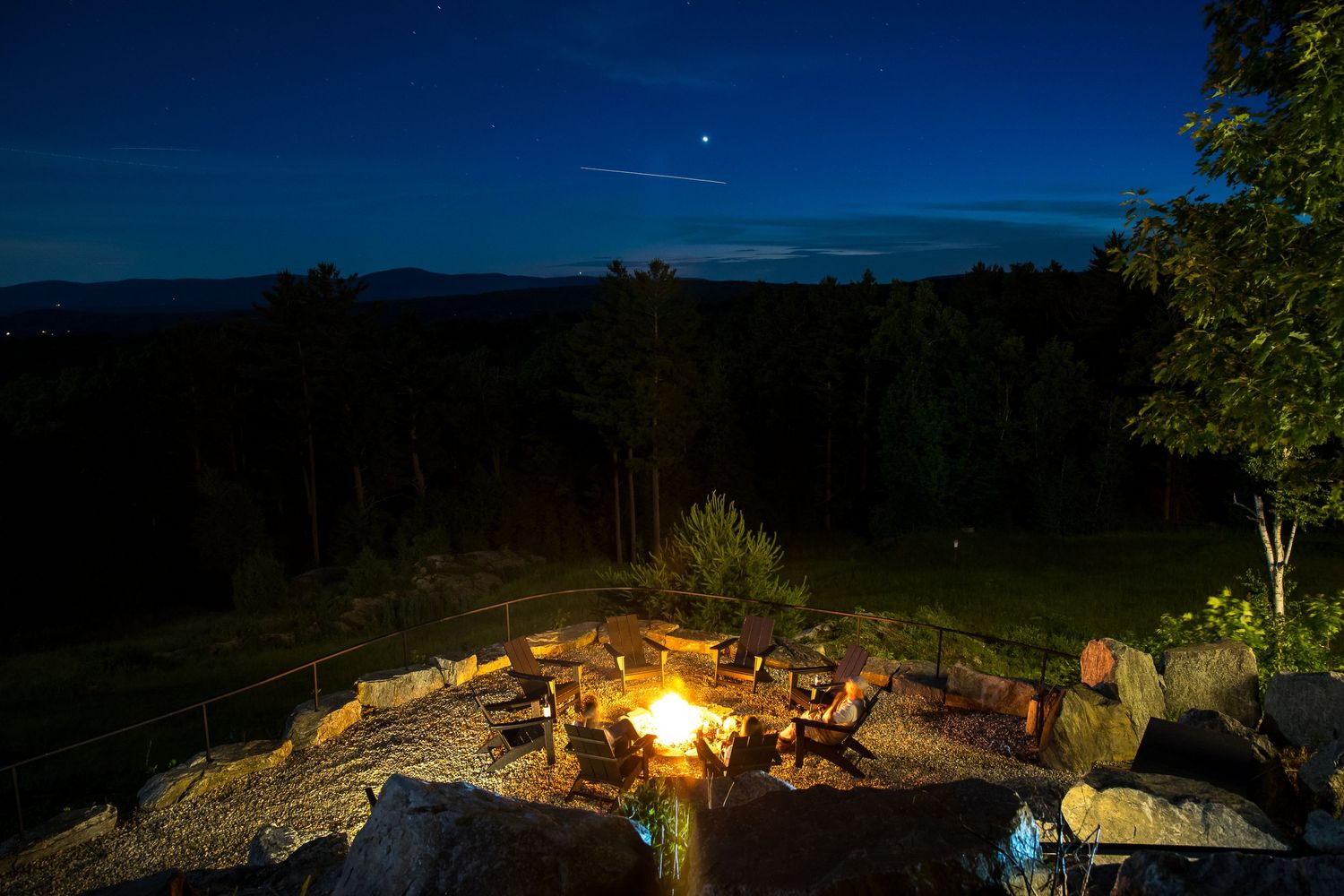 Scenic outdoor stone fire pit area with roaring flames under a starry night sky with mountain views.
