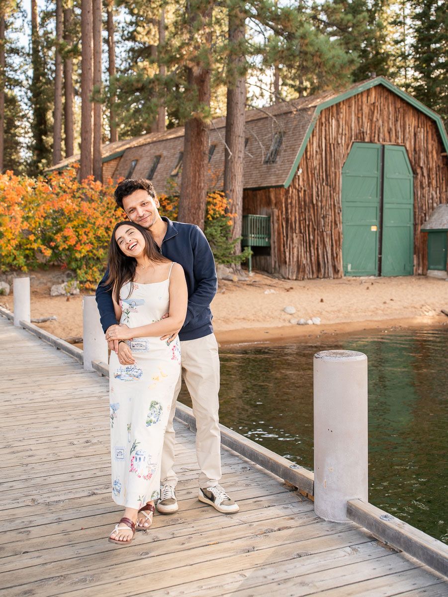 Couple embracing on a lakeside dock with a rustic boathouse and autumn foliage in the background.