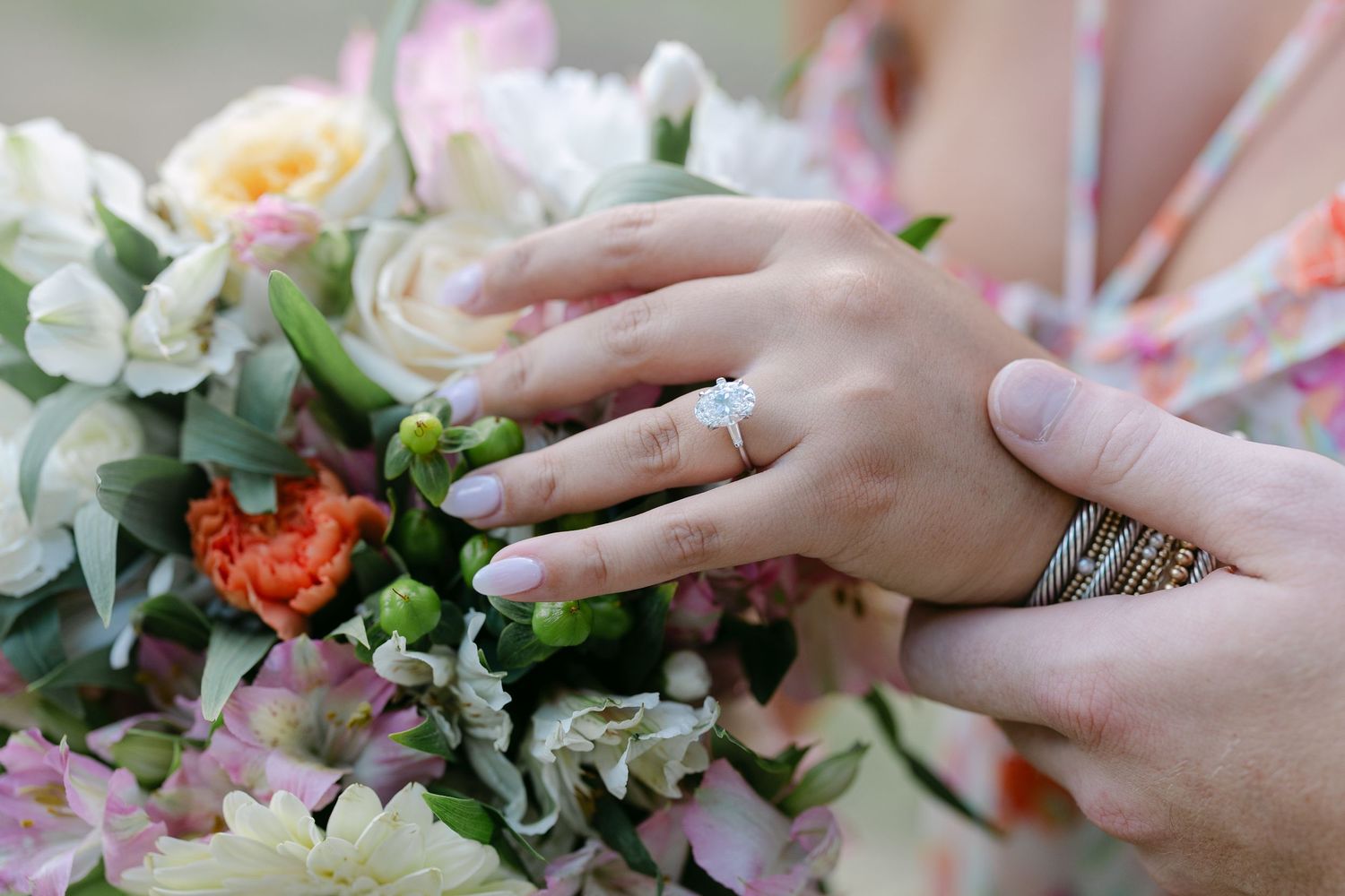 Close-up of hands with engagement ring resting on a colorful wedding bouquet with pink, white, and orange flowers.