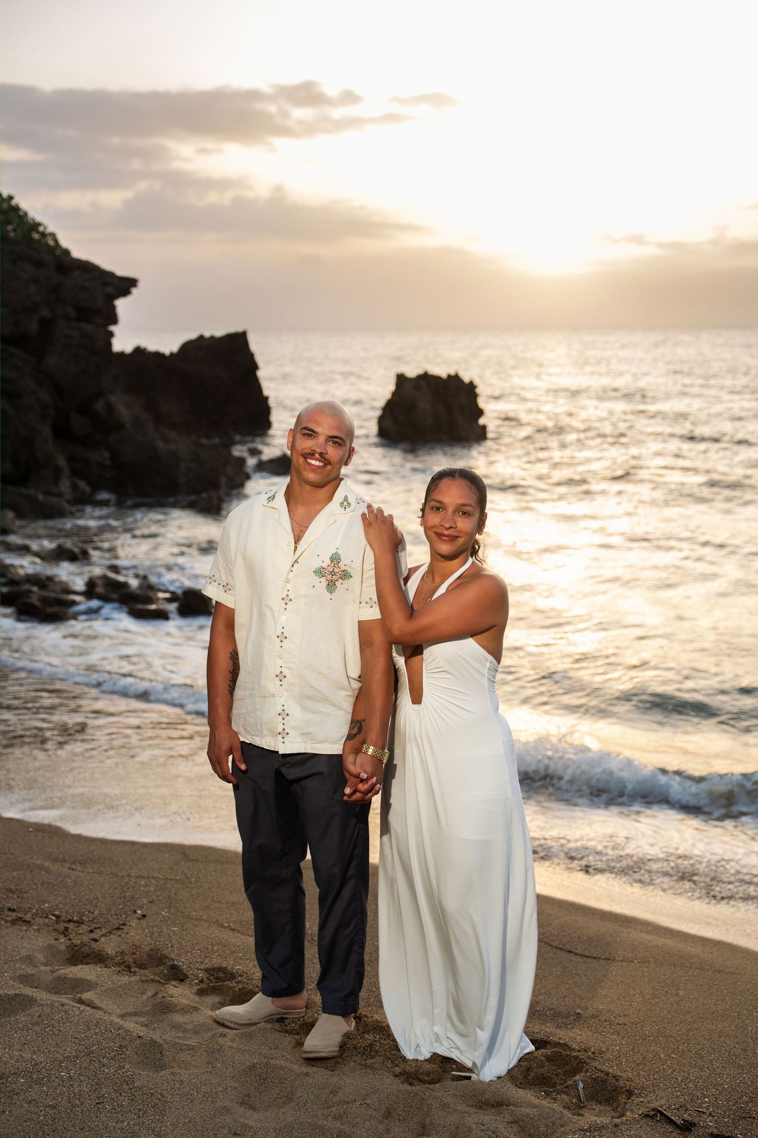 Couple posing on a sandy beach at sunset, woman in white dress, man in white shirt, with ocean waves and rocks behind them.