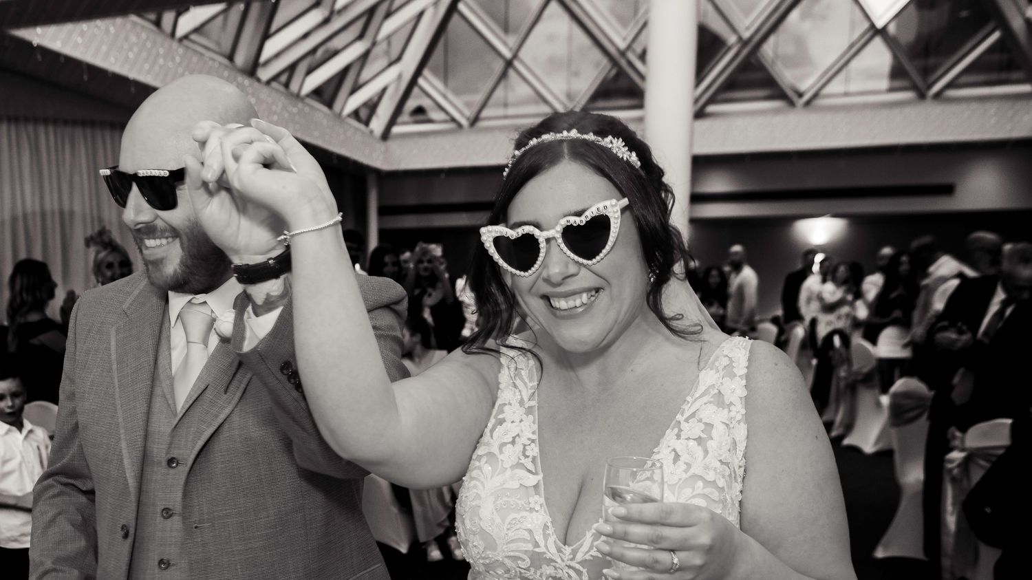 Black and white wedding photo of smiling bride in heart sunglasses and groom dancing at reception celebration.