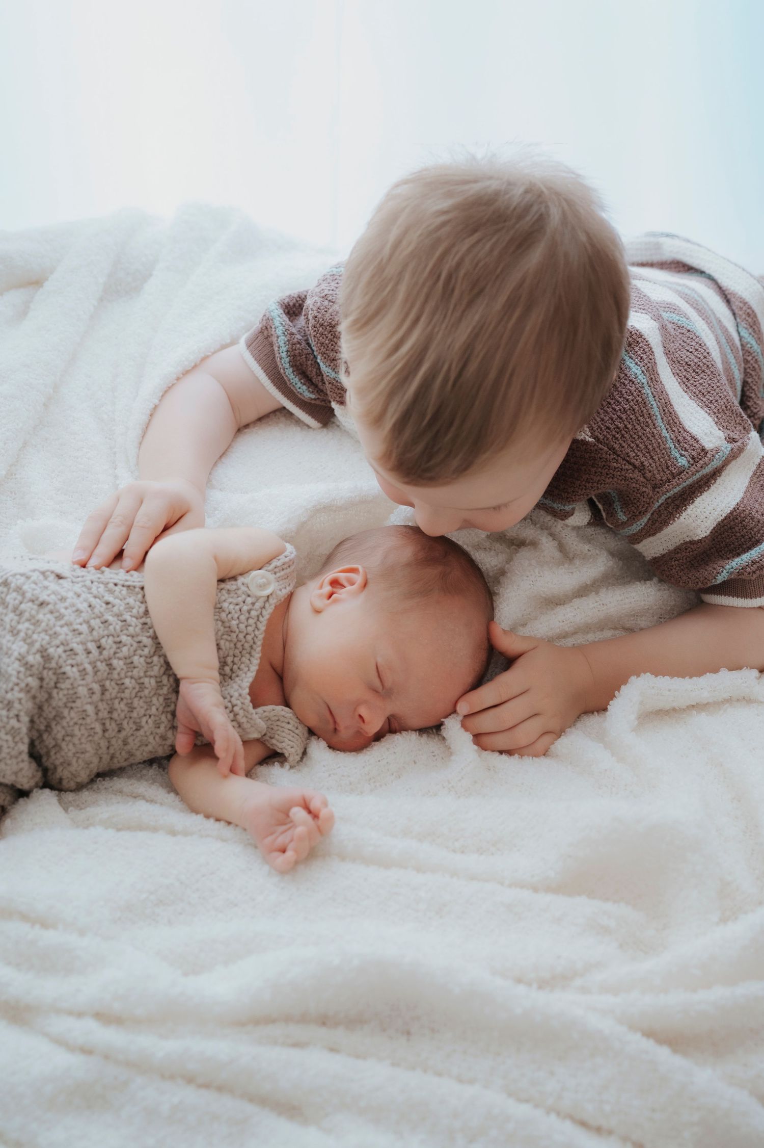 Toddler boy gently kissing sleeping newborn baby sibling on white fluffy blanket in tender moment.