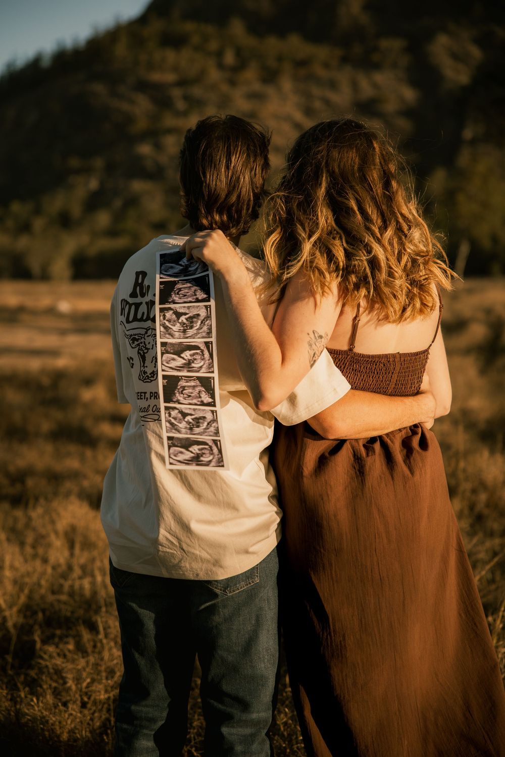 Couple embracing in golden field at sunset, man wearing ultrasound photo strip on shirt, mountains in background.