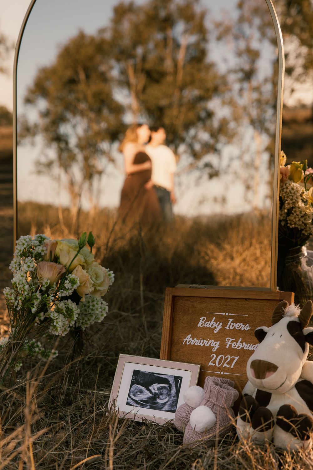 Outdoor pregnancy announcement with ultrasound photo, baby booties, cow toy, and sign reading Baby Ivers Arriving February 2027.