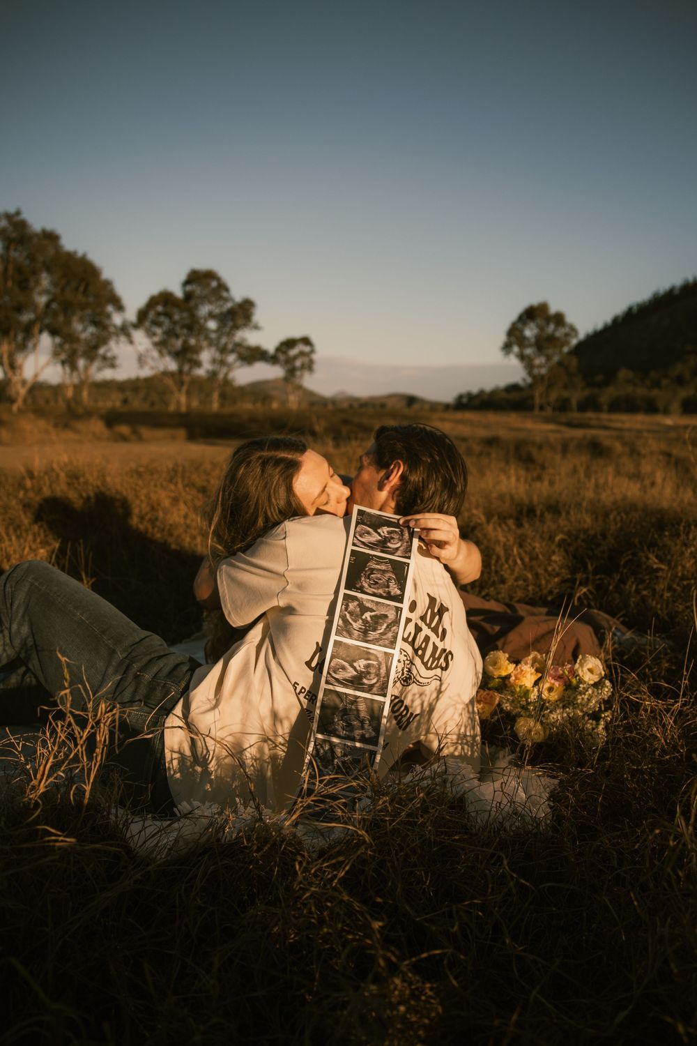 Couple kissing at sunset in a field, holding ultrasound photos, announcing pregnancy outdoors.
