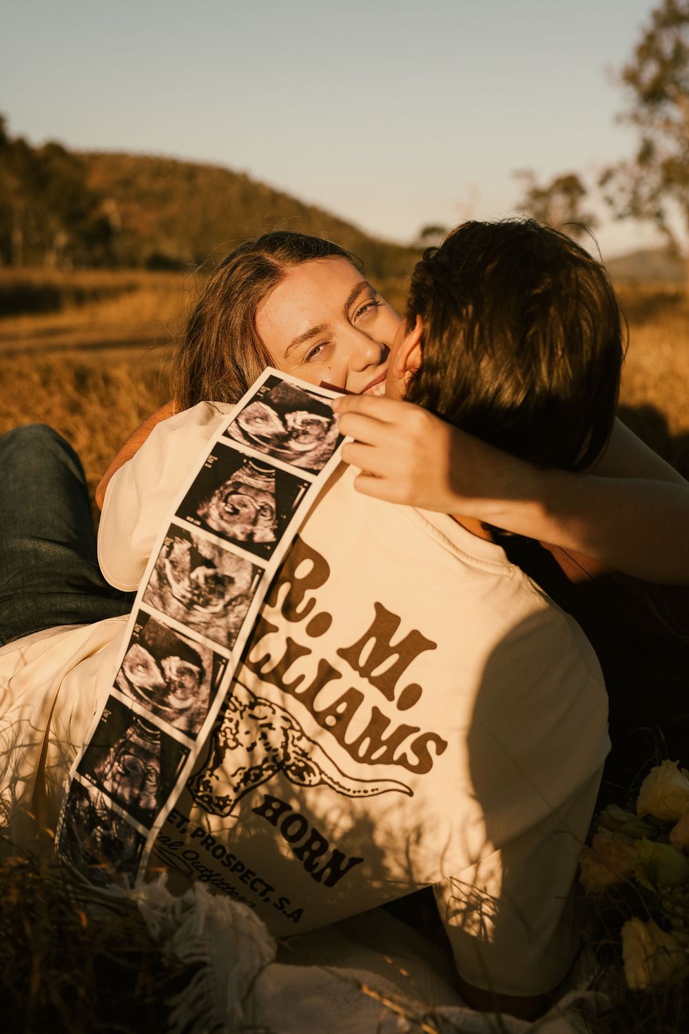 Happy couple sharing a kiss outdoors at golden hour, woman smiling and holding ultrasound photos announcing pregnancy.