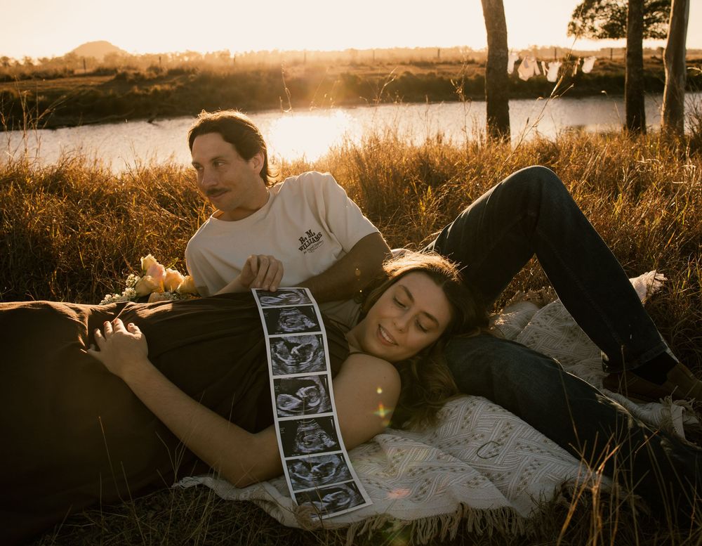 Couple lying in golden hour field, woman resting on mans lap holding ultrasound strip, pregnancy announcement photo.