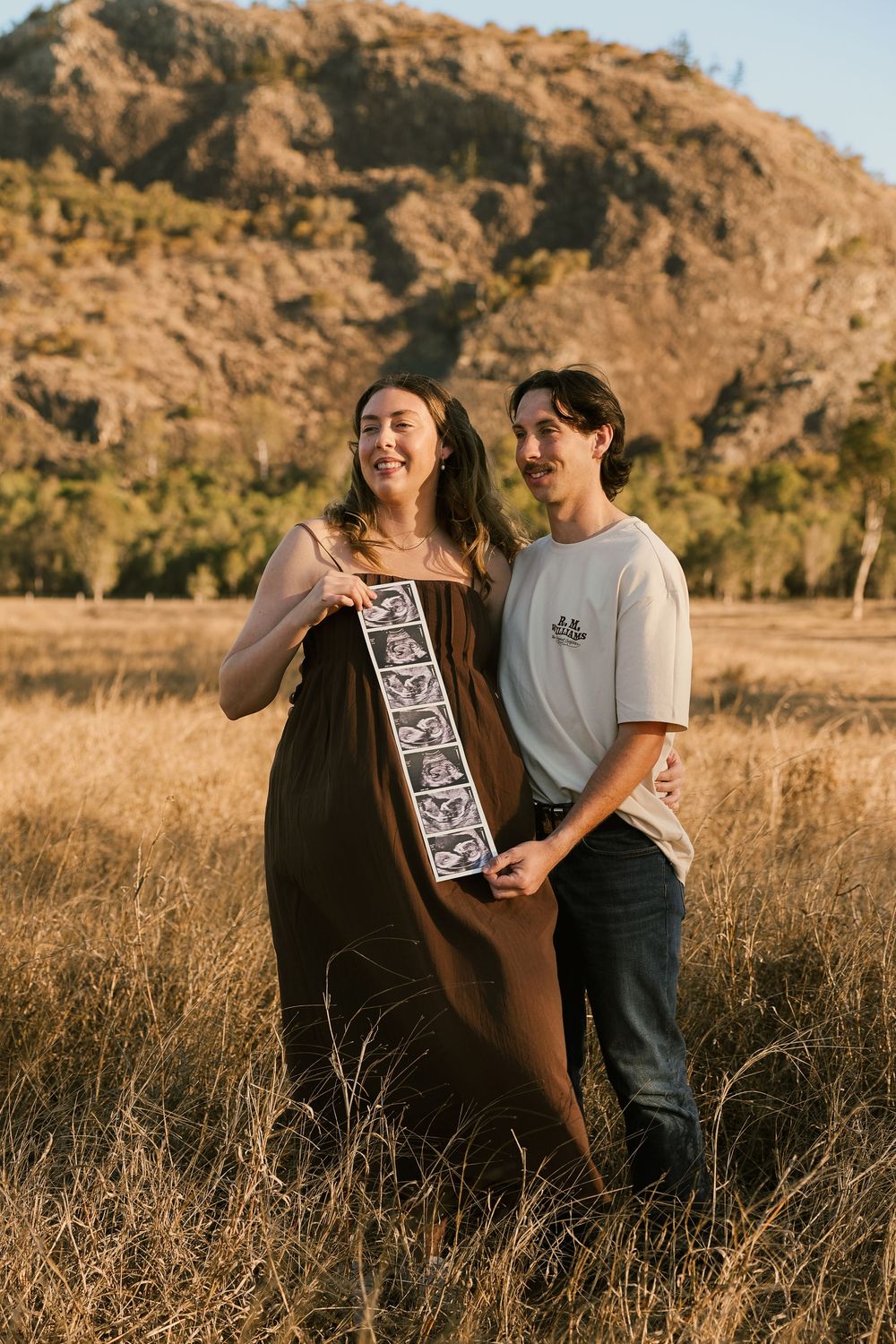 Couple holding ultrasound photos in golden grass field with rocky hill backdrop during maternity photo shoot.