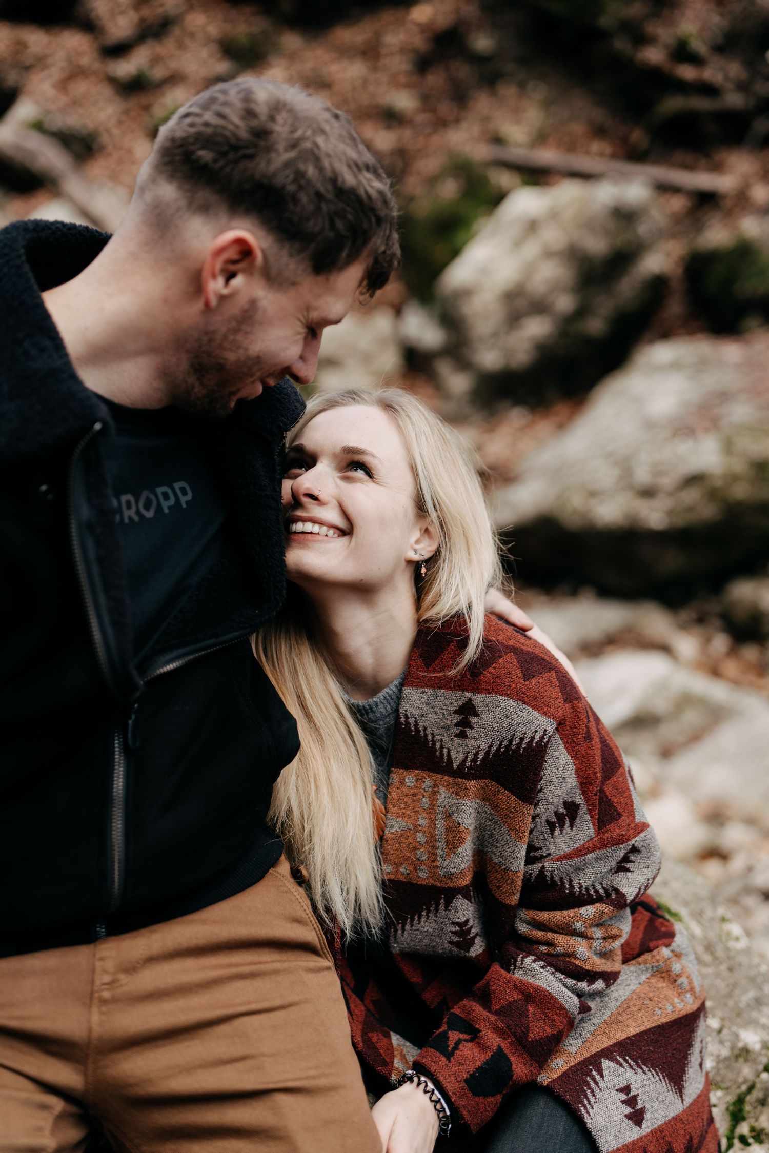 A smiling blonde woman gazes up at a man in a forest setting, both wearing cozy sweaters in an outdoor couples portrait.