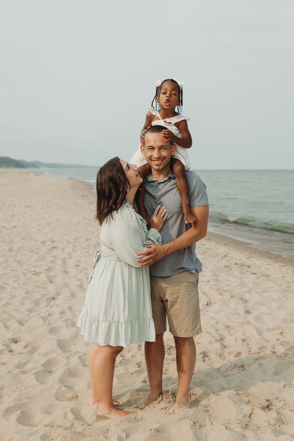 Happy family of three enjoying a sunny beach day, with child sitting on father's shoulders while mother embraces them.