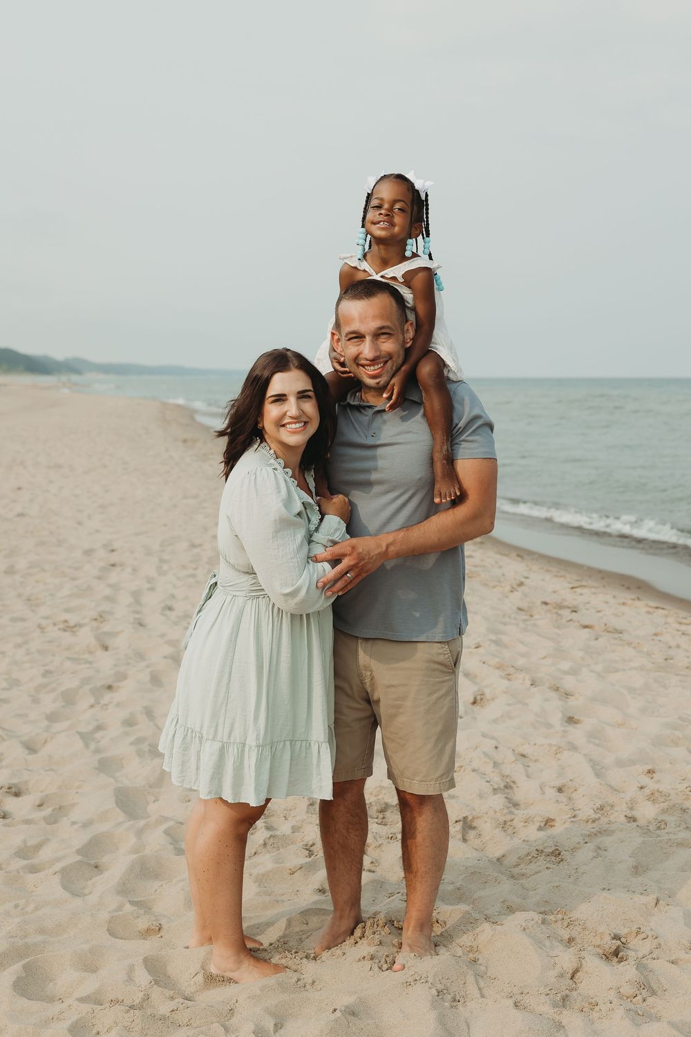 Happy family of three posing on a sandy beach, with parents smiling and child sitting on dad's shoulders.