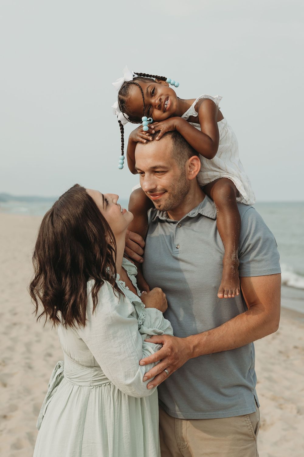 Happy interracial family of three enjoying a beach day, with parents smiling at their young daughter sitting on dad's shoulders.