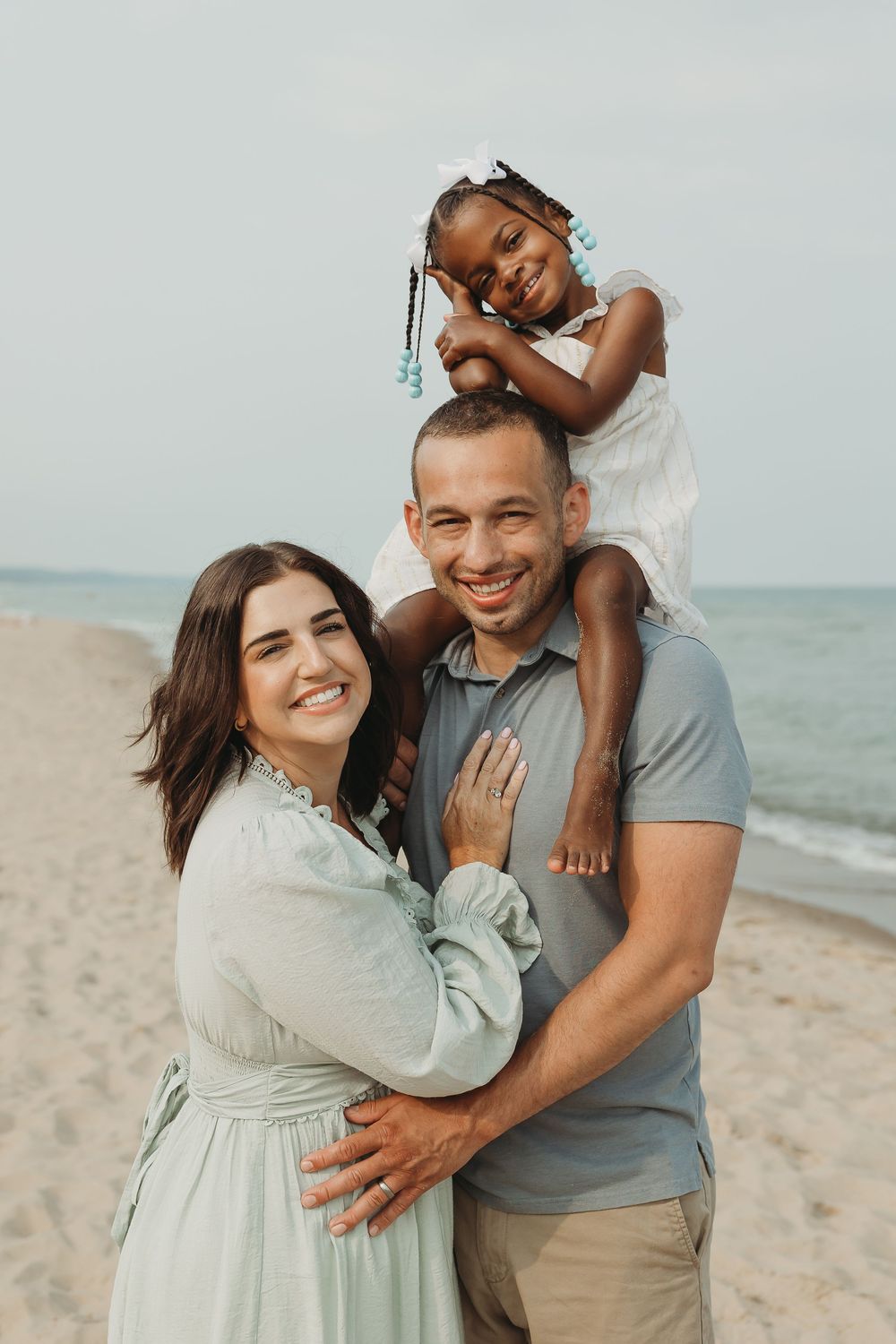 Happy interracial family of three posing on a sandy beach, with a young girl sitting on her father's shoulders.