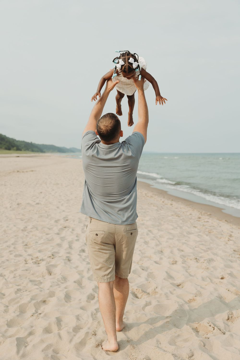 Father lifting baby joyfully on sandy beach with ocean waves and clear sky in background.