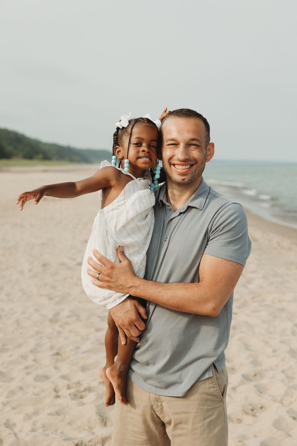Smiling man holds young girl on sandy beach, waves and trees visible in background.