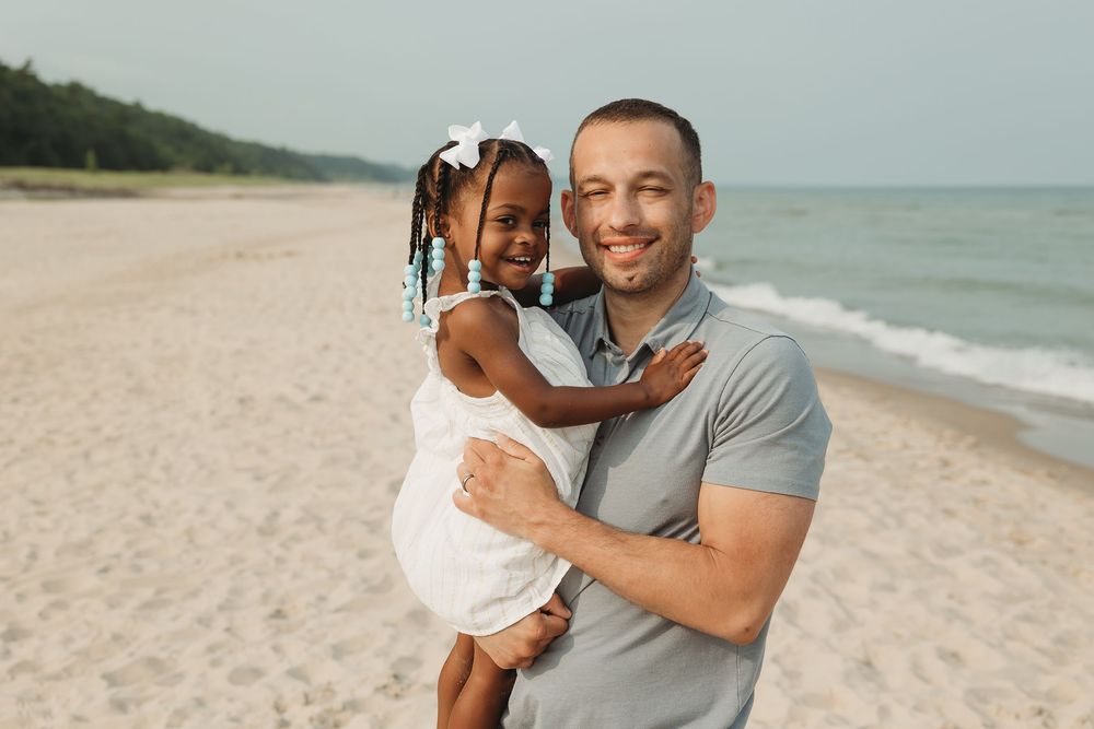 A smiling man holds a young girl in a white dress on a sandy beach near the water.