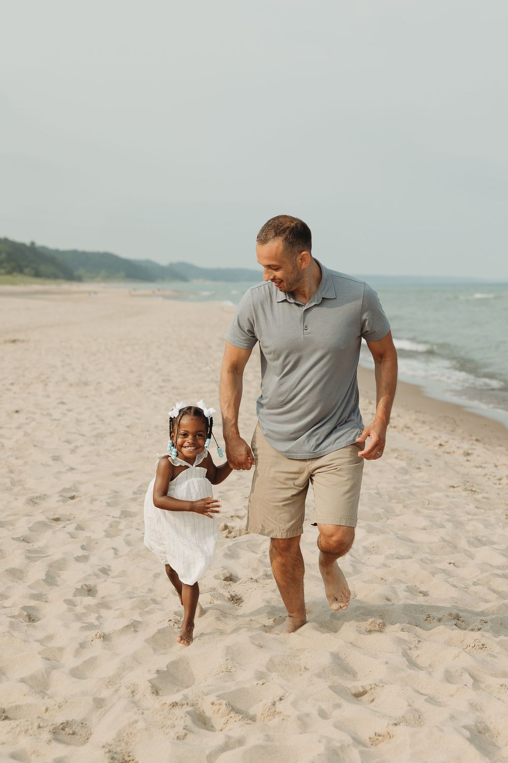 A man and young girl running and laughing together on a sandy beach near the ocean on a cloudy day.
