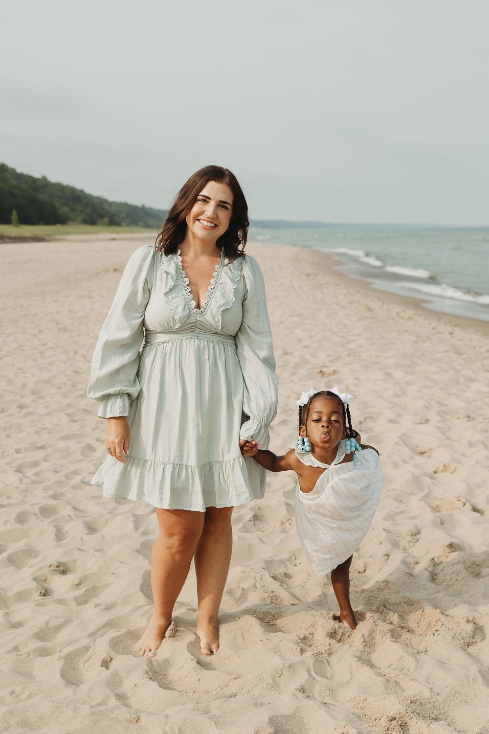 A smiling woman in a mint dress holds hands with a young girl in white on a sandy beach.