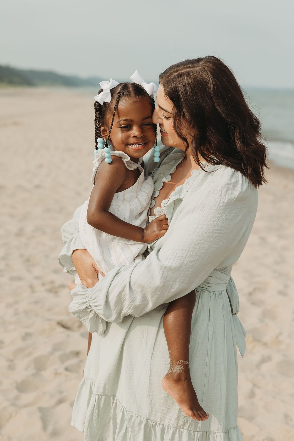 A woman in a mint dress holds a smiling girl in white on a sandy beach, sharing a tender moment.
