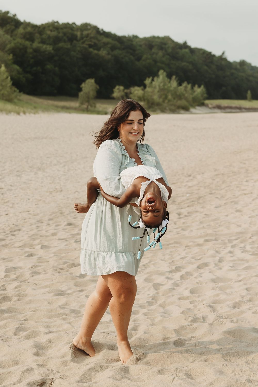 A smiling woman in a mint dress playfully holds a laughing child upside down on a sandy beach.