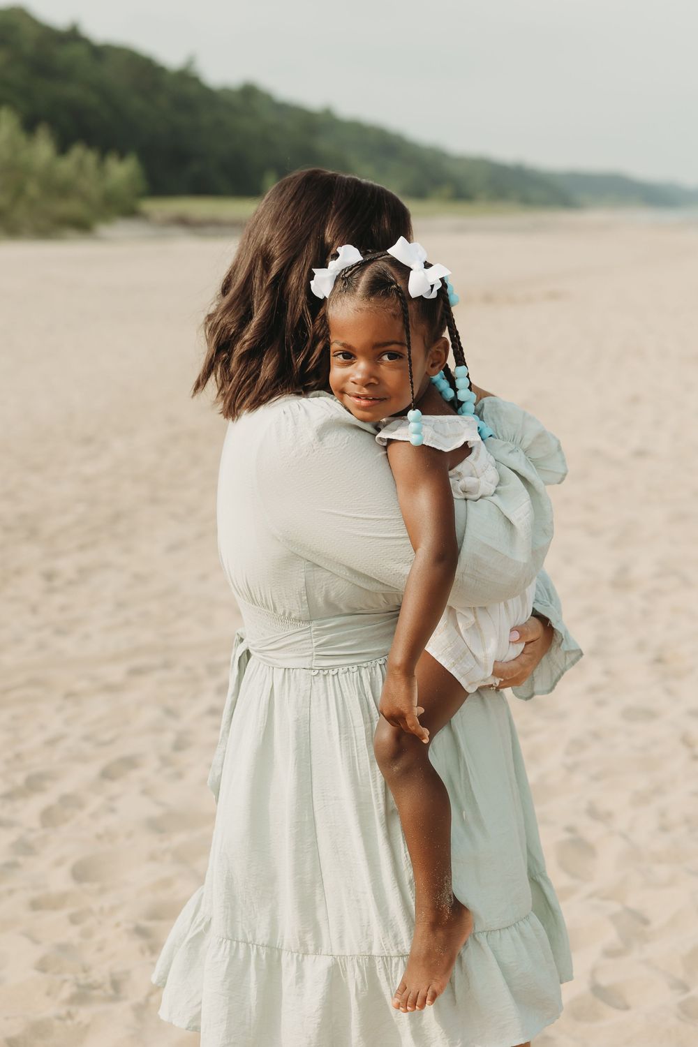 A woman in a mint dress holds a young girl with white bows on a sandy beach.