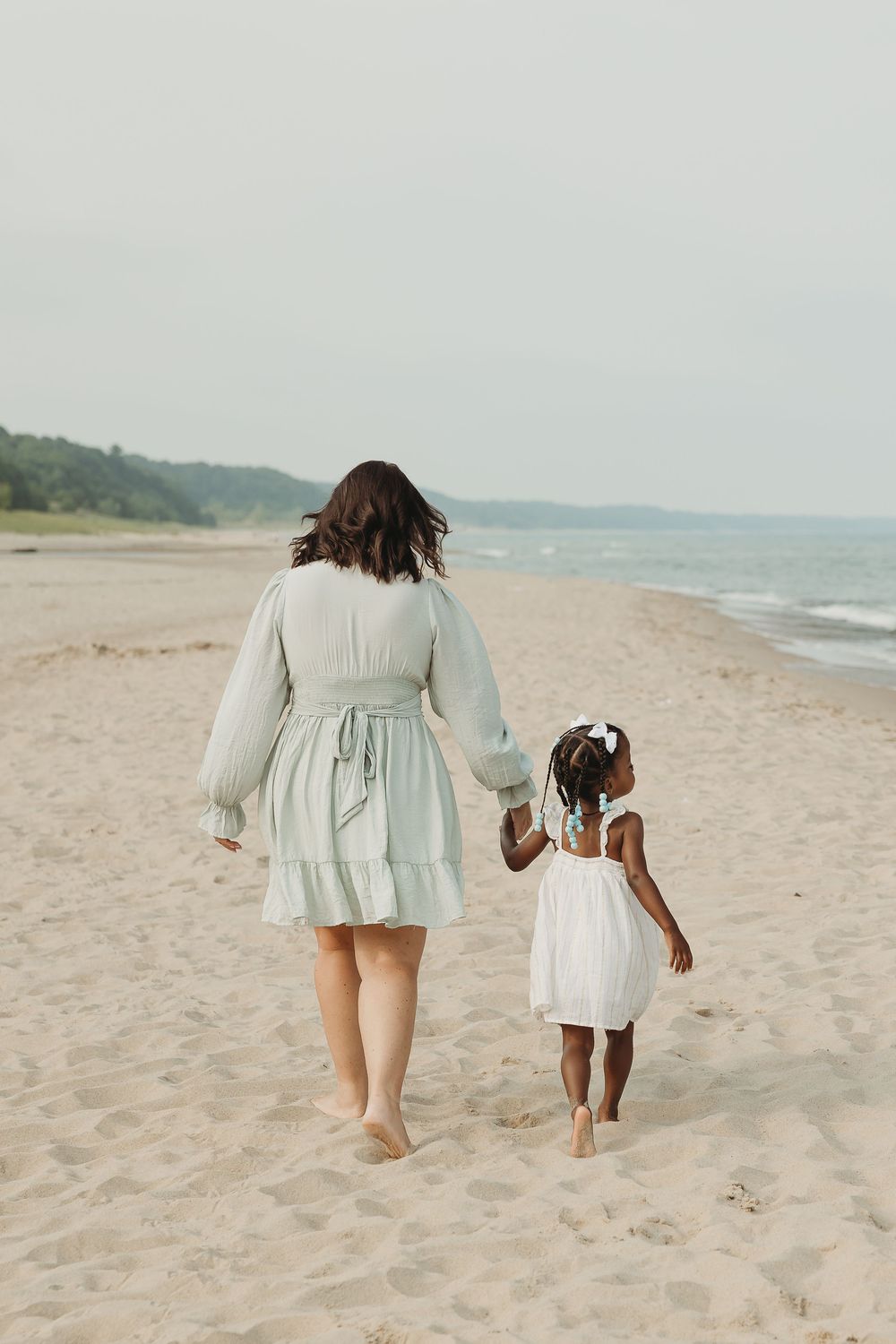 A woman and young girl walk hand-in-hand along a sandy beach, both wearing light dresses, viewed from behind.