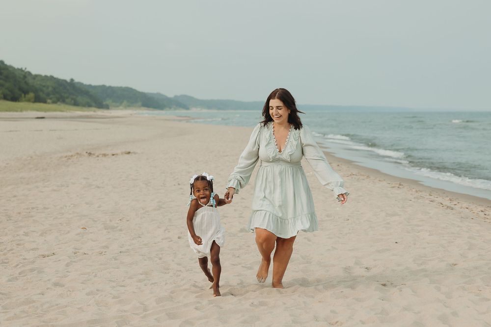 Woman in mint dress holding hands with young girl running on sandy beach by the ocean.