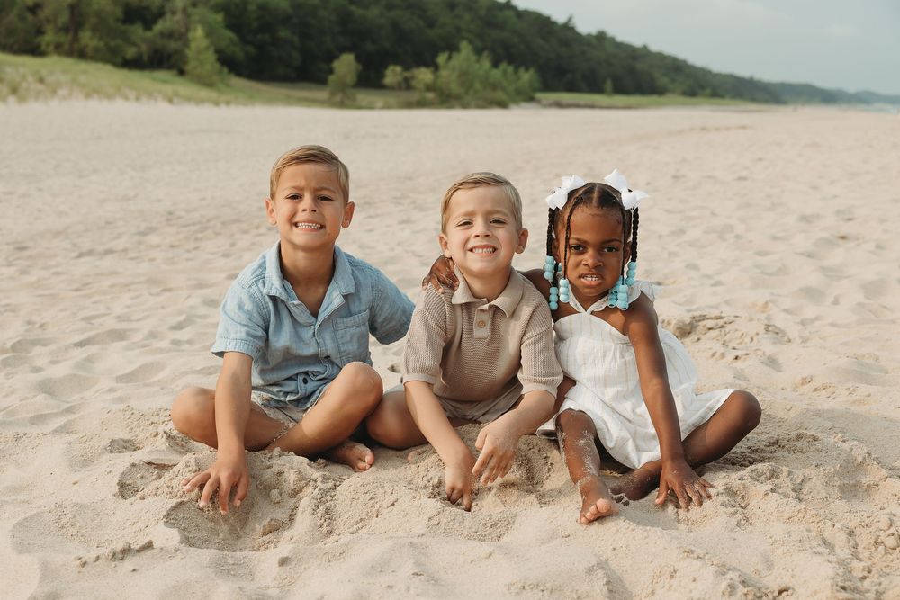 Three happy children sitting together on a sandy beach, smiling for a photo on a sunny day.
